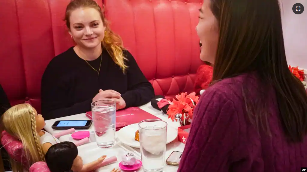 Marisa Dragos, 23, left, along with her childhood friend Lisa Costantino, 24, right, sit for lunch at American Girl Cafe with two store-borrowed dolls, Friday, Dec. 2, 2022, in New York. (AP Photo/Bebeto Matthews)