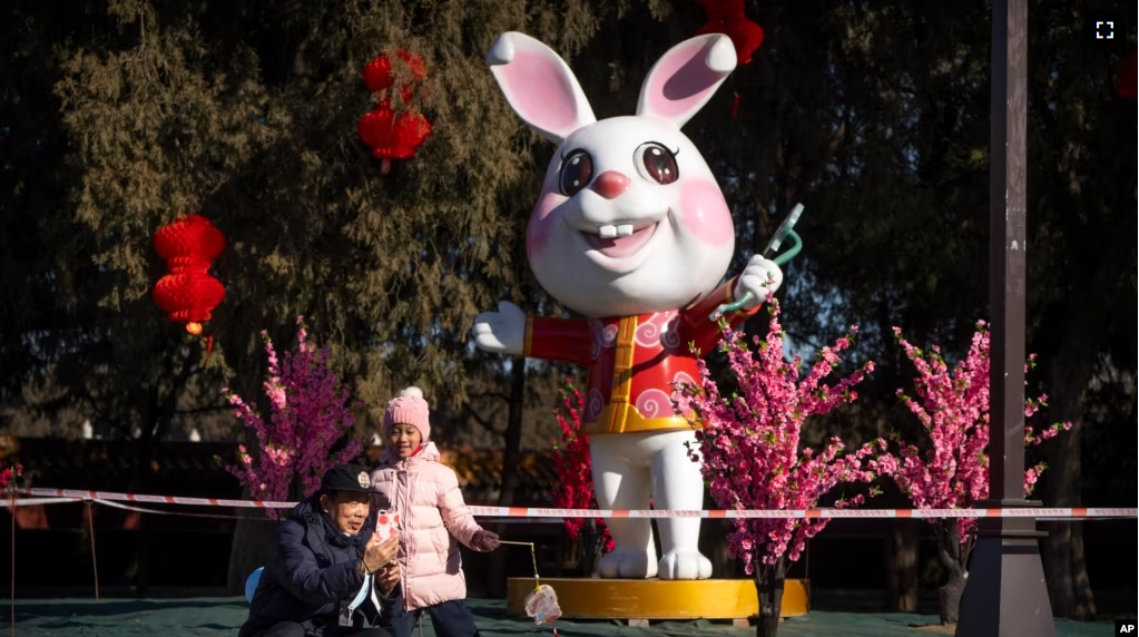 A man and girl pose for a selfie in front a of a statue of a rabbit for the upcoming Lunar New Year at a public park in Beijing, Friday, Jan. 20, 2023. (AP Photo/Mark Schiefelbein)