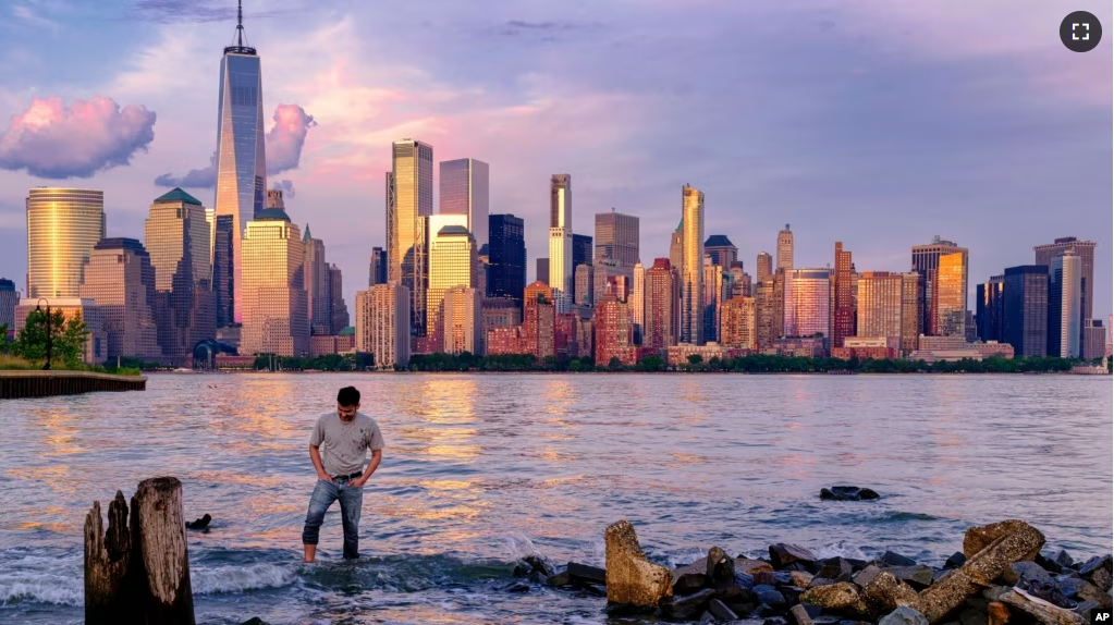 FILE — A man wades through a canal in Jersey City as the sun sets on the lower Manhattan skyline of New York City, May 31, 2022. (AP Photo/J. David Ake)