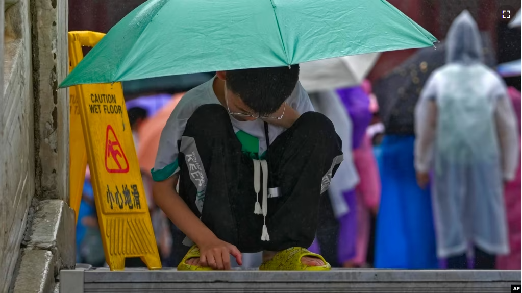 FILE - A boy covering himself with an umbrella from the rain, browses a smartphone placed on the ground at the Forbidden City in Beijing on July 13, 2023. (AP Photo/Andy Wong)