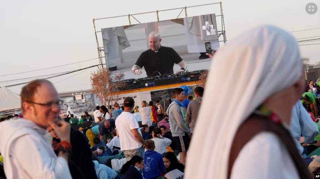 FILE - Roman Catholic priest Guilherme Peixoto, on video screen, plays techno music to help pilgrims wake up at Parque Tejo in Lisbon on Aug. 6, 2023, where Pope Francis will preside over a mass celebrating the 37th World Youth Day later in the day. (AP Photo/Ana Brigida)